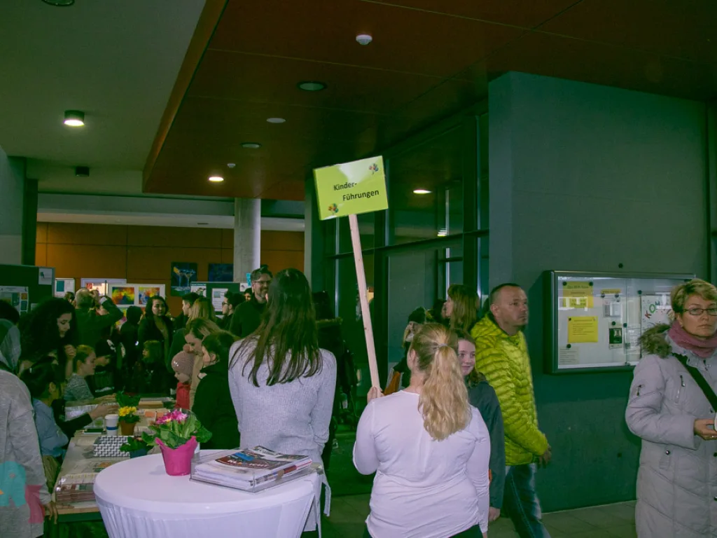 Besucher an Stehtischen im Foyer, dahinter ein Wegweiser.